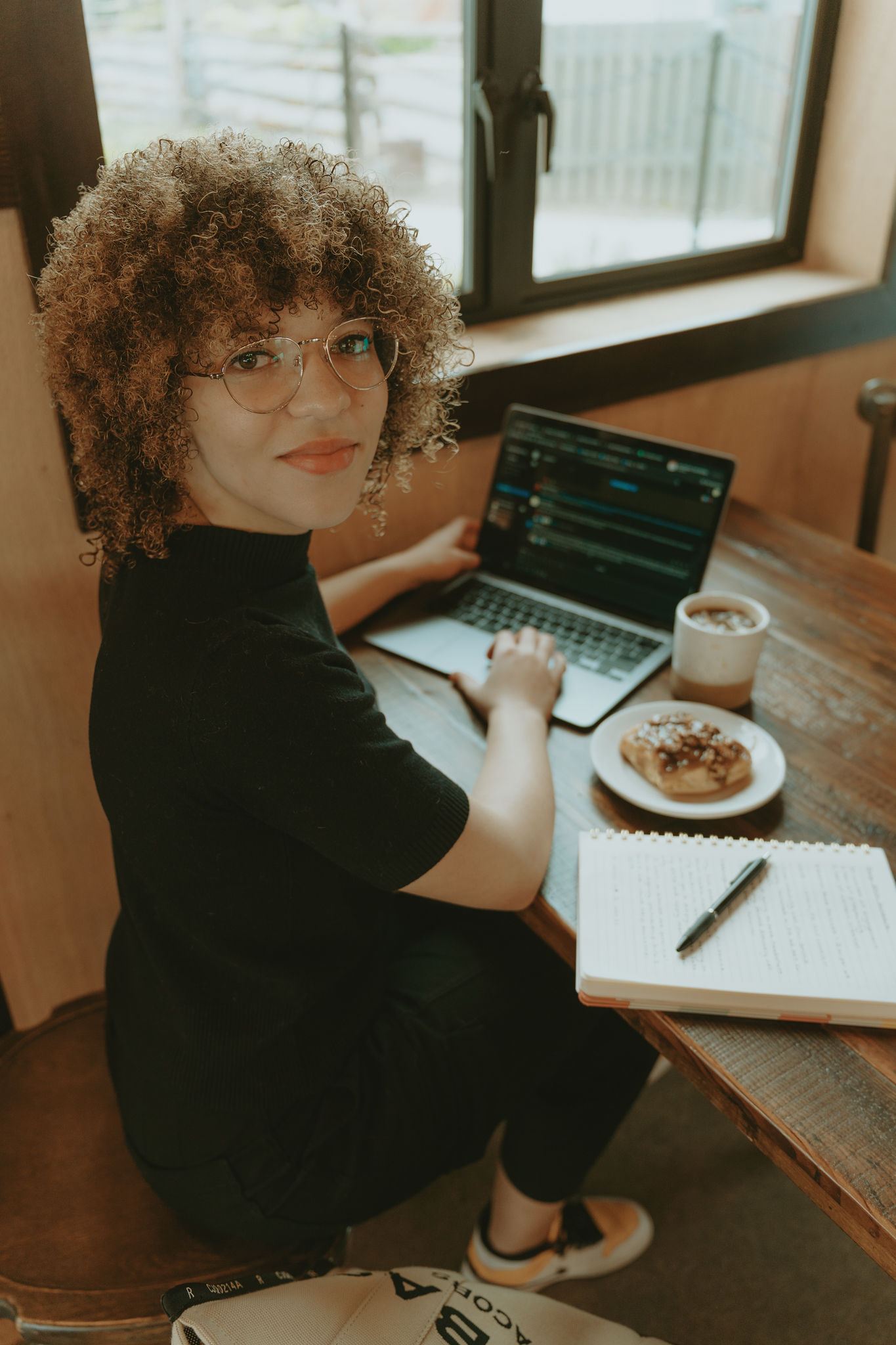 Shaylee is sitting at a table in a cafe. She is smiling to camera as she types on her laptop with a coffee and sticky bun on the table beside her.